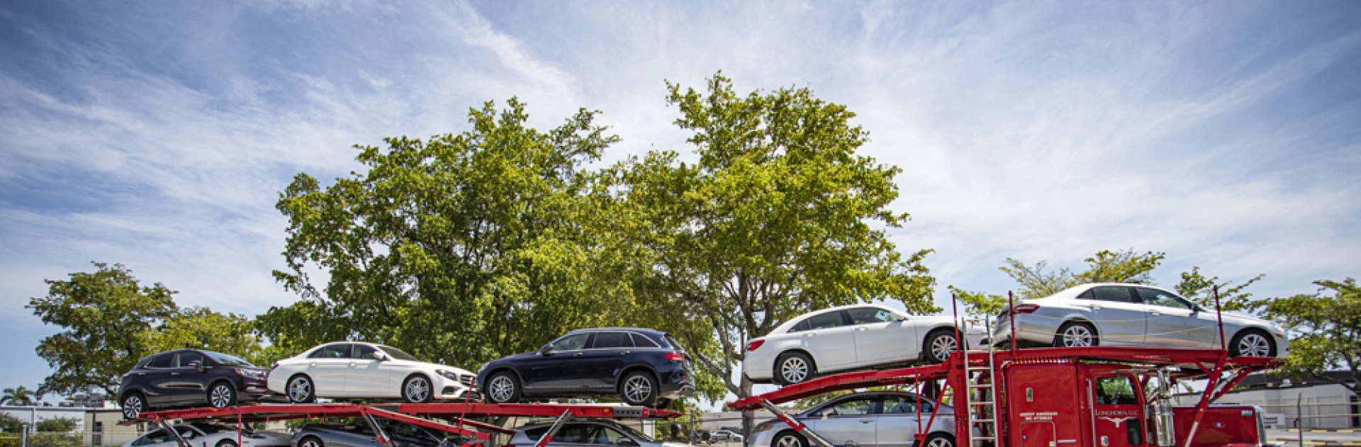 Red car carrier loaded with sedans parked under trees, hero background image