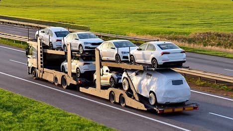 Open carrier auto transport Open car carrier hauling white sedans with one wrapped vehicle on a highway through green fields